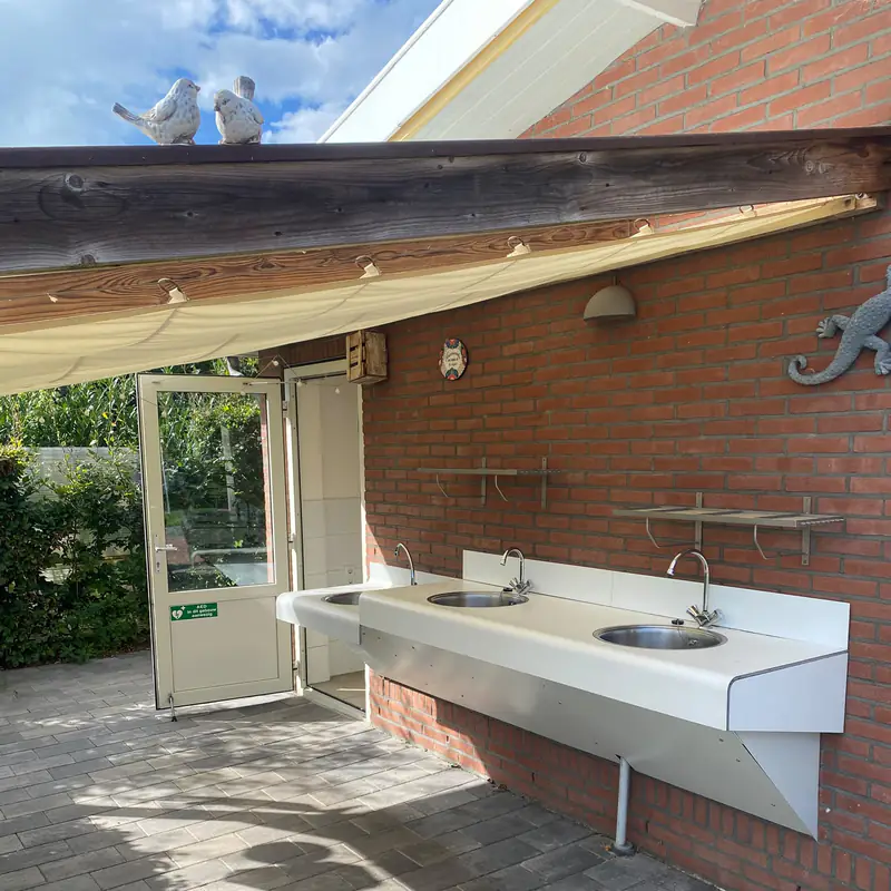 De afwasplek buiten onder de overkapping, met drie spoelbakken en aanrechtblad langs de muur