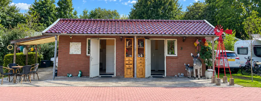 Het sanitairgebouw van buiten, met open deuren onder de overkapping en een terras met tafeltjes ernaast