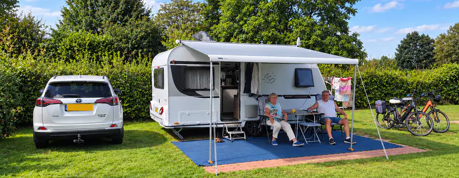 Een caravan met uitgeschoven luifel op een comfortplaats, met de auto ernaast en gasten aan tafel