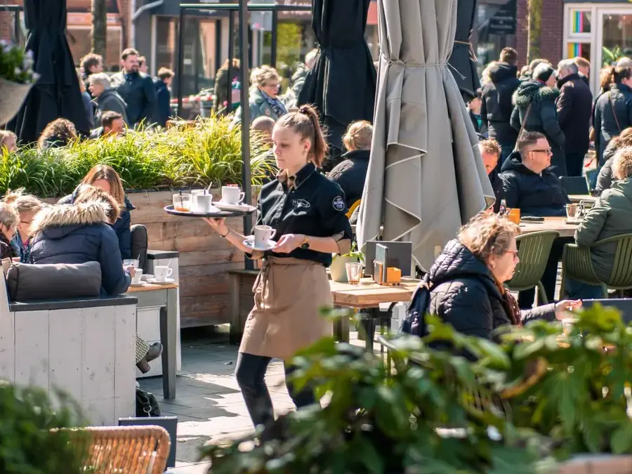 Een terras op een dorpsplein in de zon, met gasten aan de tafels en een serveerster met een dienblad