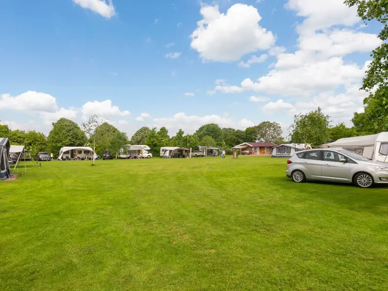Het open kampeerveld met caravans en voortenten langs de randen onder een blauwe lucht